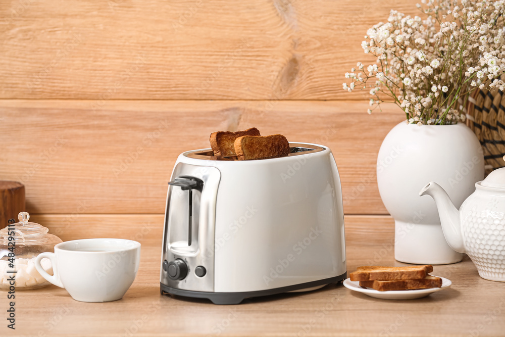 White toaster with bread slices, cup and vase on table near wooden wall