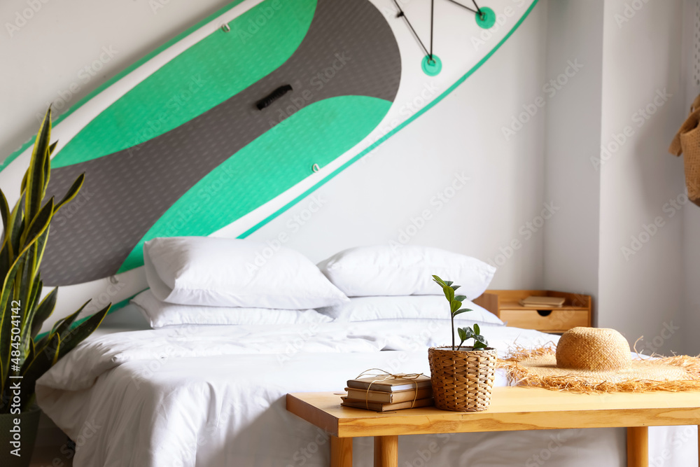 Houseplant, books and straw hat on table in bedroom interior with sup surfing board