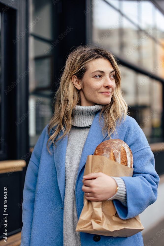 Attractive blonde woman, looking to the side, carrying bread, smiling.