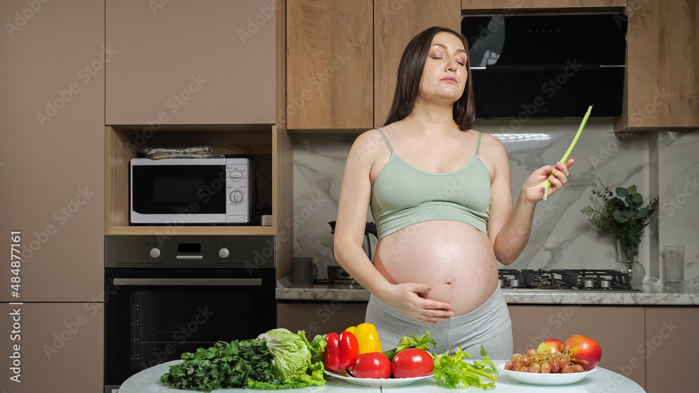 Pregnant brunette woman wearing top and training pants eats peeled celery looking at vegetables and 