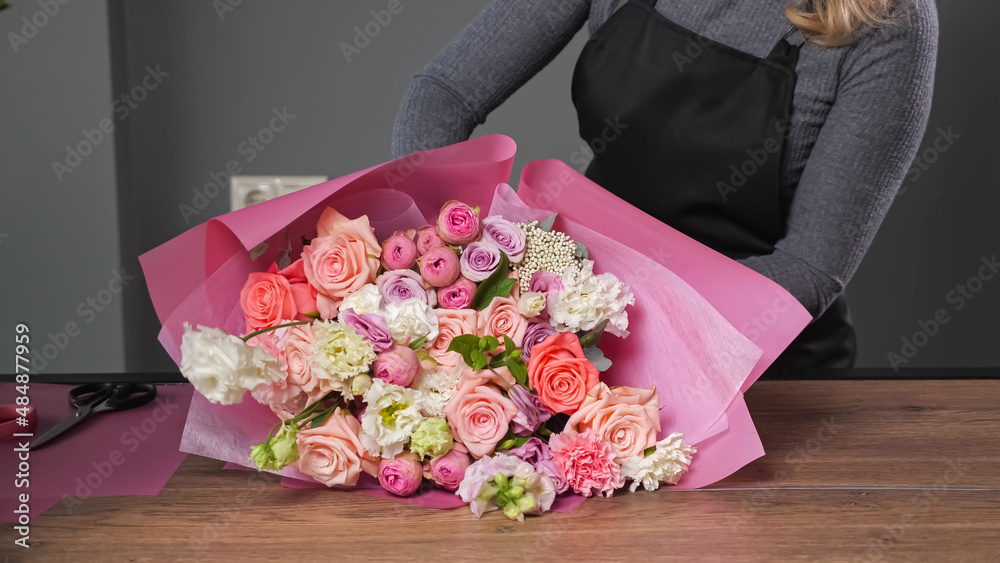 Florist woman in black apron makes elegant wedding bouquet with colorful roses wrapping in pink pape