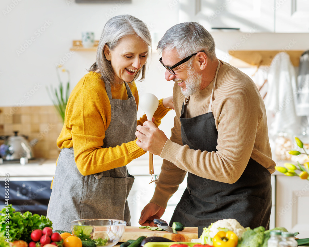 Joyful elderly couple have fun dancing and singing while cooking together in kitchen