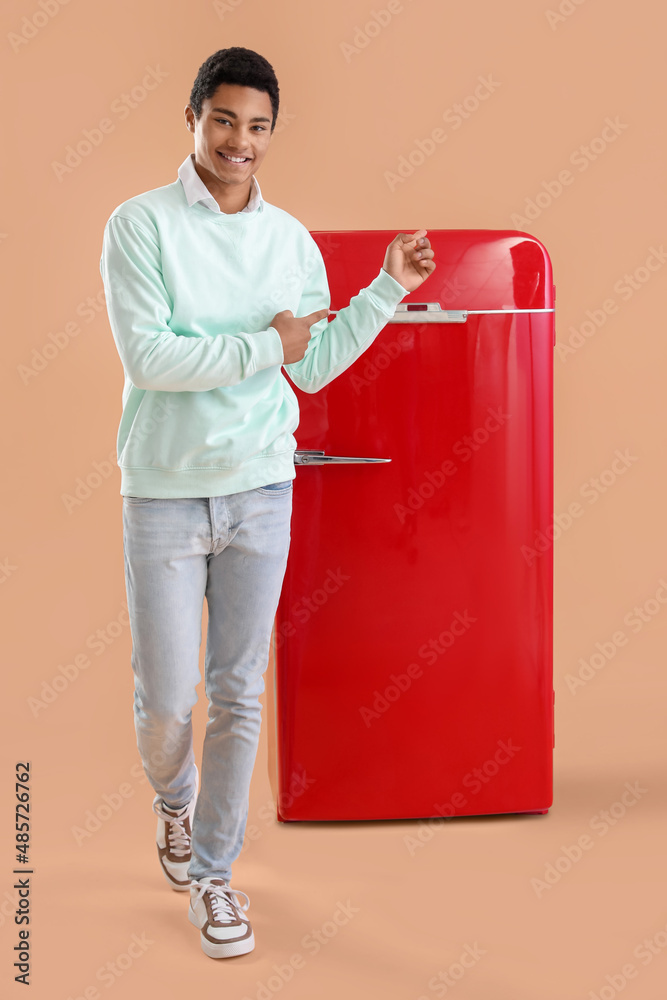 Smiling African-American man pointing at stylish red fridge on color background