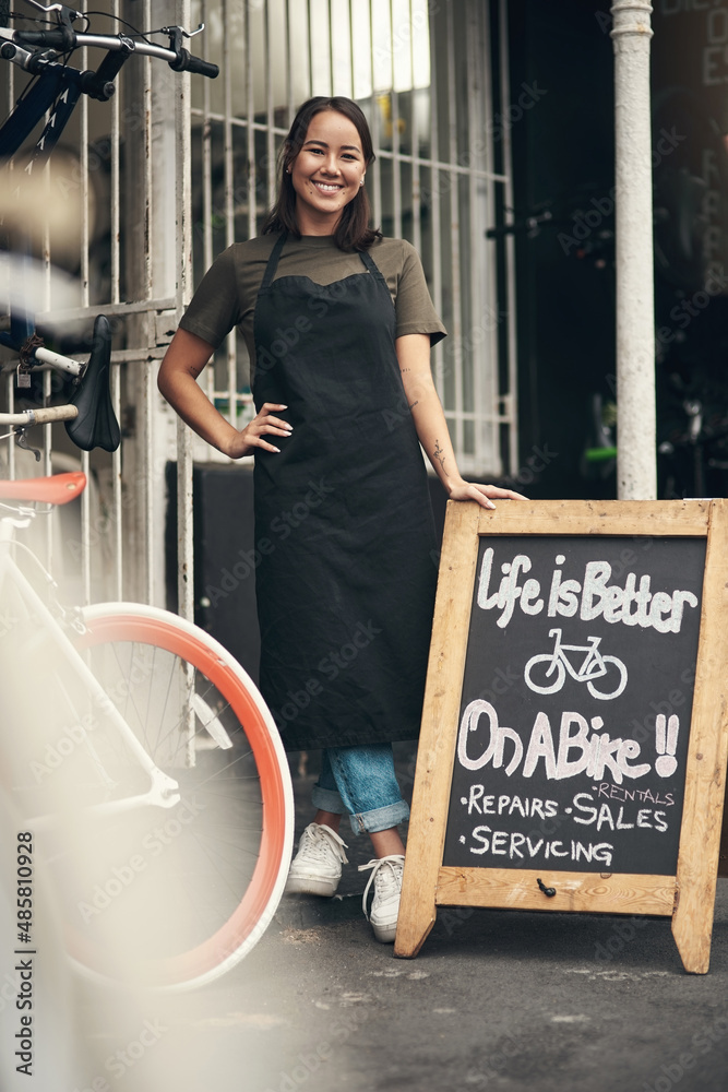 Come on in. Full length shot of an attractive young woman standing outside her bicycle shop during t