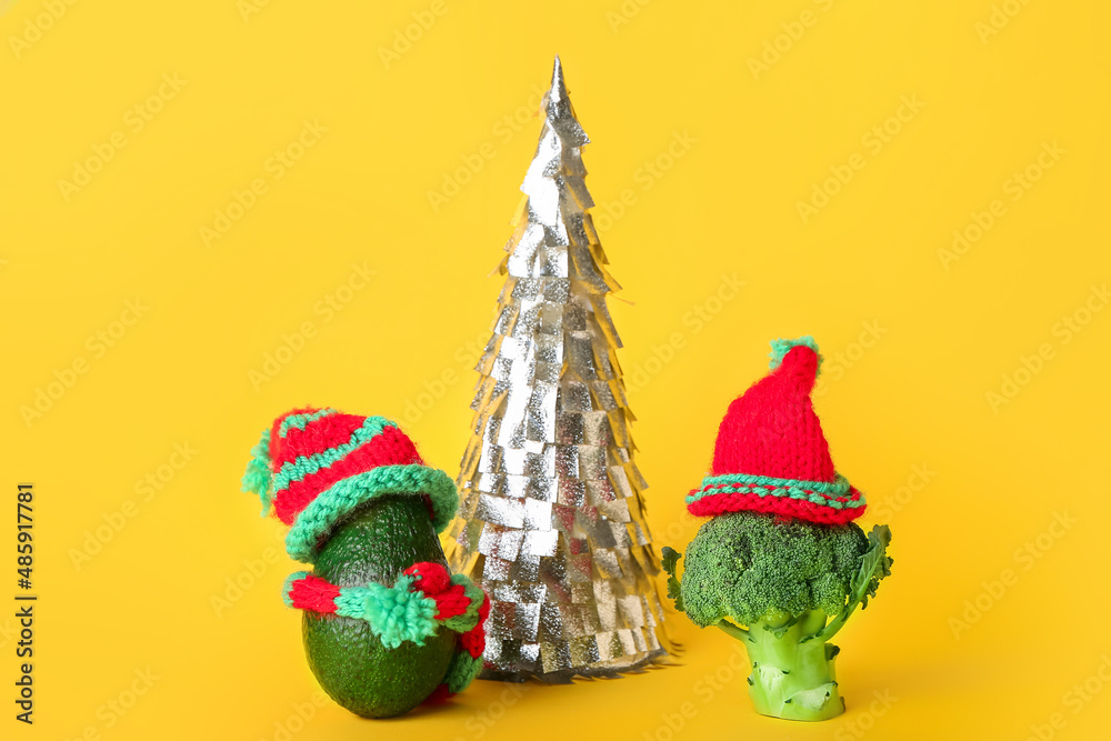 Composition with Christmas tree, avocado and broccoli in knitted hats on yellow background