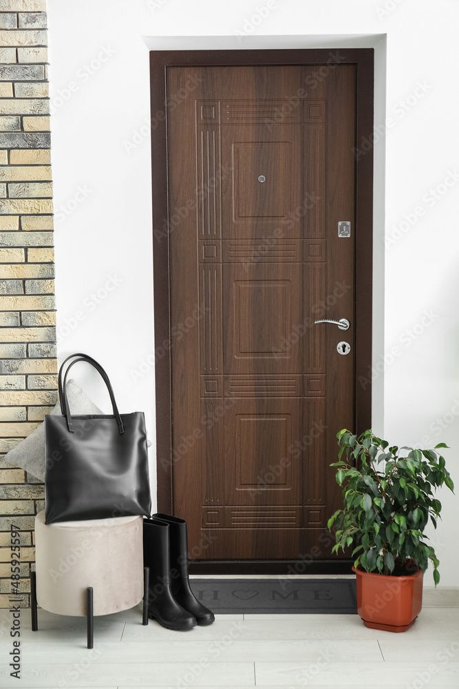 Pouf with bag, gumboots, houseplant and black mat near dark wooden door