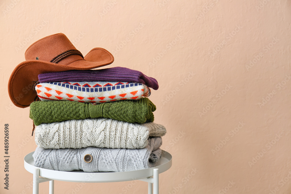 Stack of sweaters and stylish hat on table near color wall