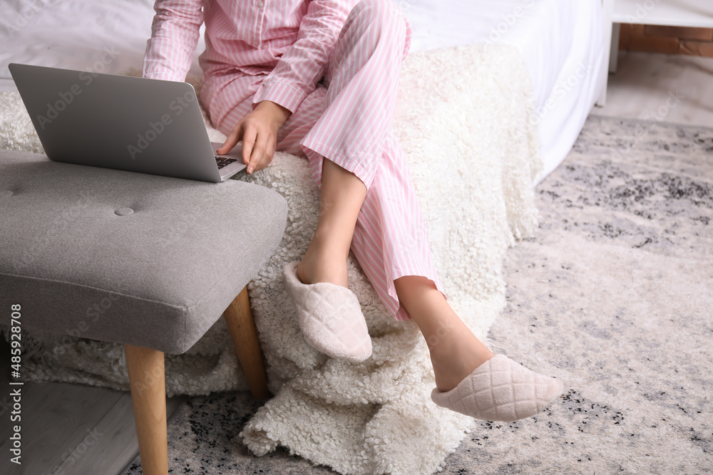 Young woman in soft comfortable slippers using laptop while sitting on bed