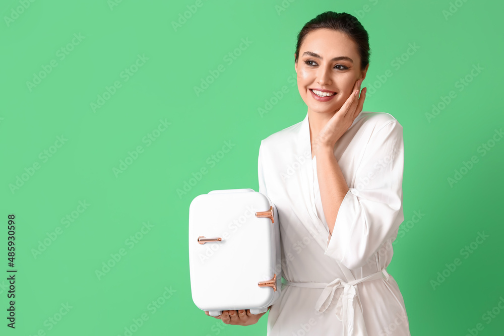 Happy young woman with small cosmetic refrigerator on green background