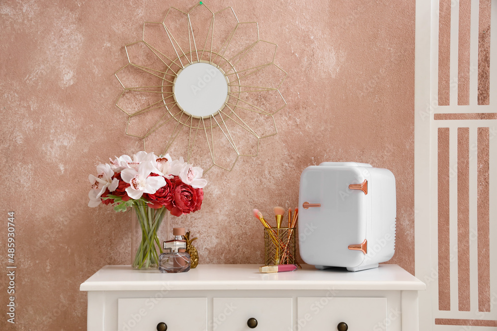 Small cosmetic refrigerator, makeup brushes and vase with flowers on table near color wall