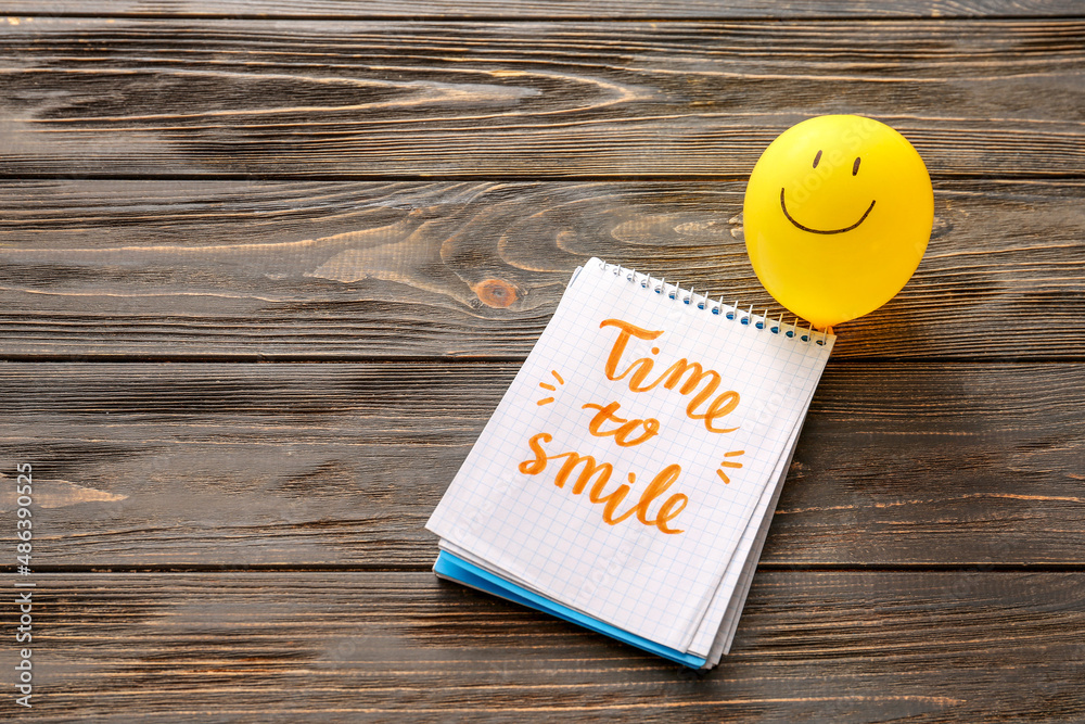 Air balloon with drawn happy face and notebook with text TIME TO SMILE on table