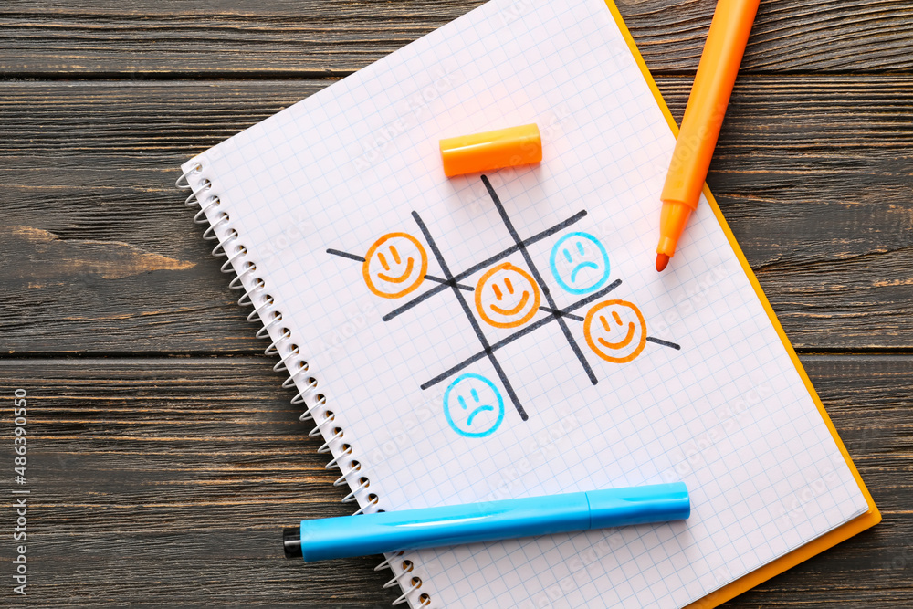 Notebook with tick-tack-toe game and drawn happy and sad faces on table