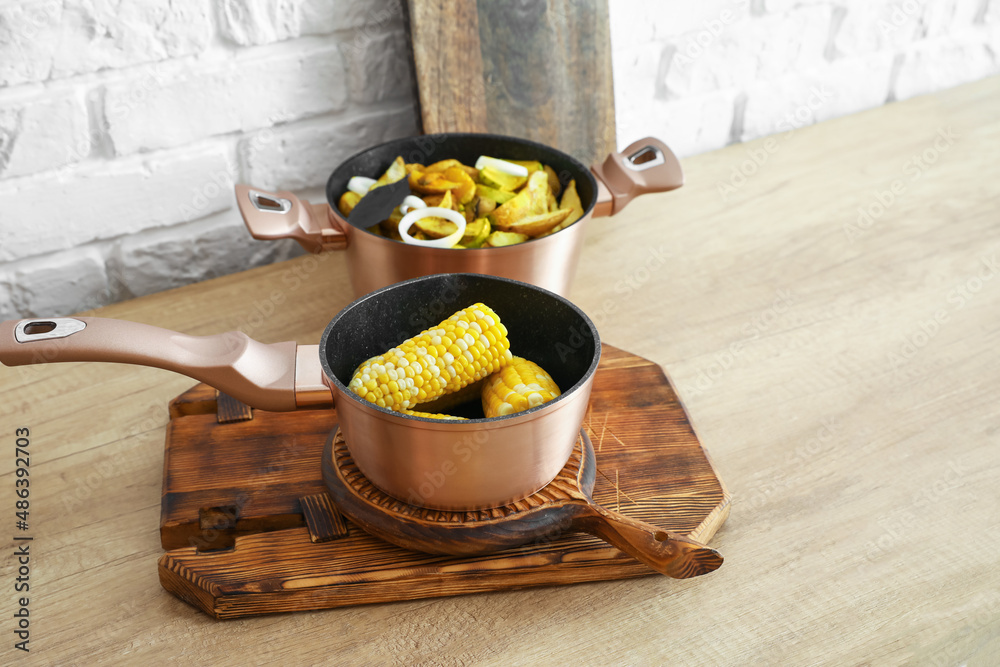 Sauce pan with corn cobs on wooden board and cooking pot with vegetable stew on table in kitchen