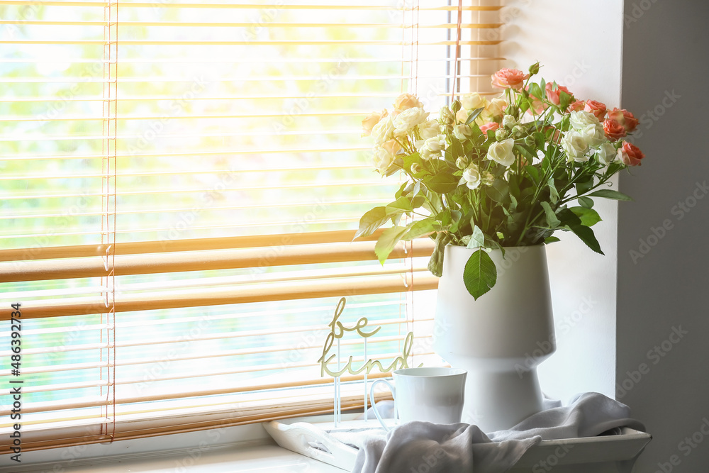 Vase with beautiful fresh roses and cup of coffee on windowsill