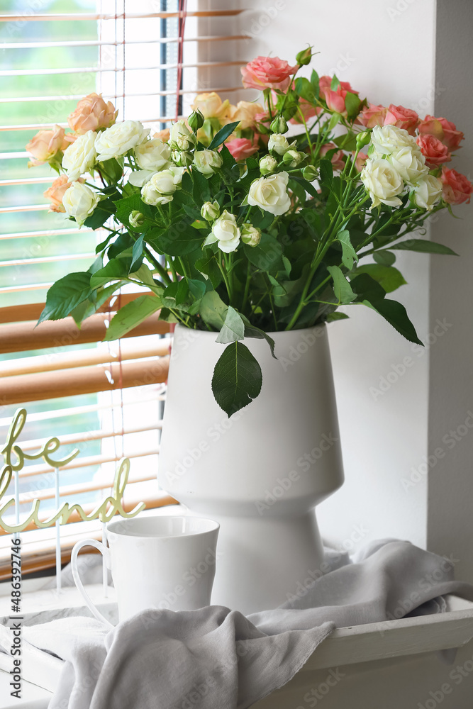 Vase with beautiful fresh roses and cup of coffee on windowsill