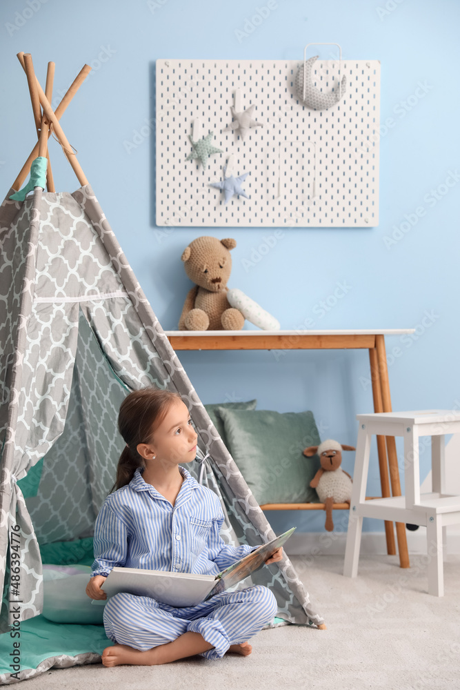 Cute little girl in pajamas reading book near play tent at home