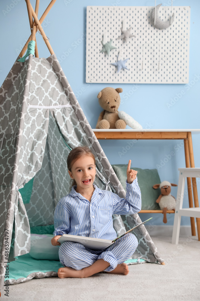 Cute little girl in pajamas with book pointing at hanging peg board on blue wall