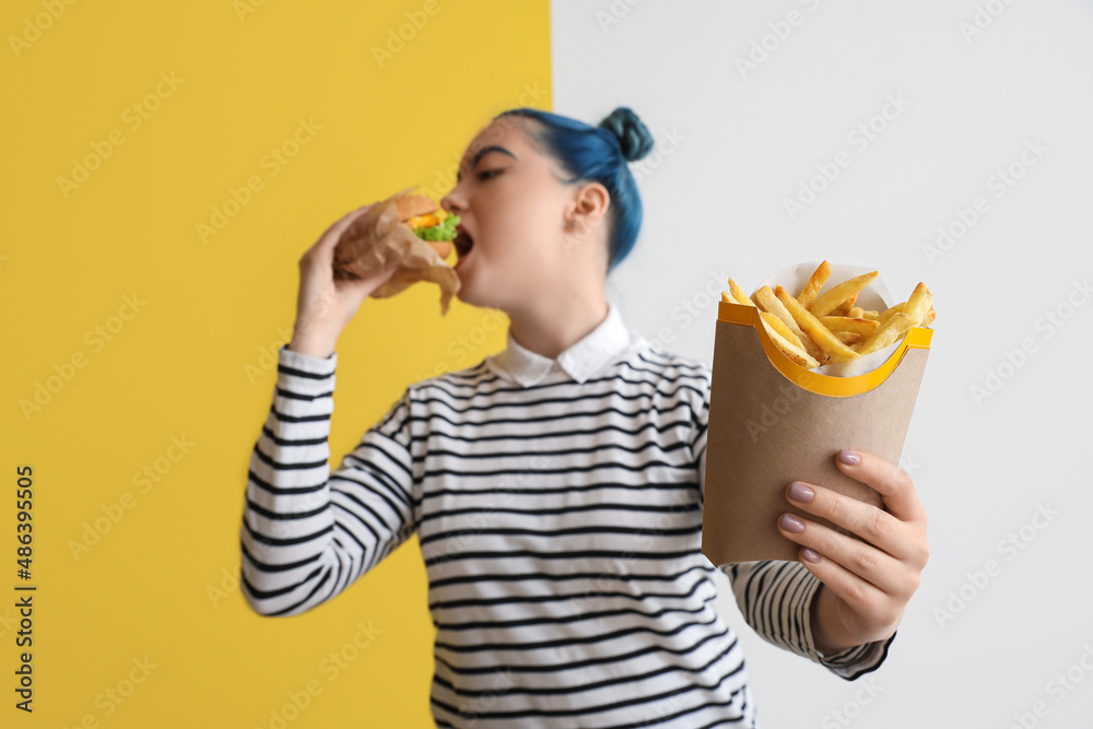 Young woman with unusual hair eating burger and french fries on color background