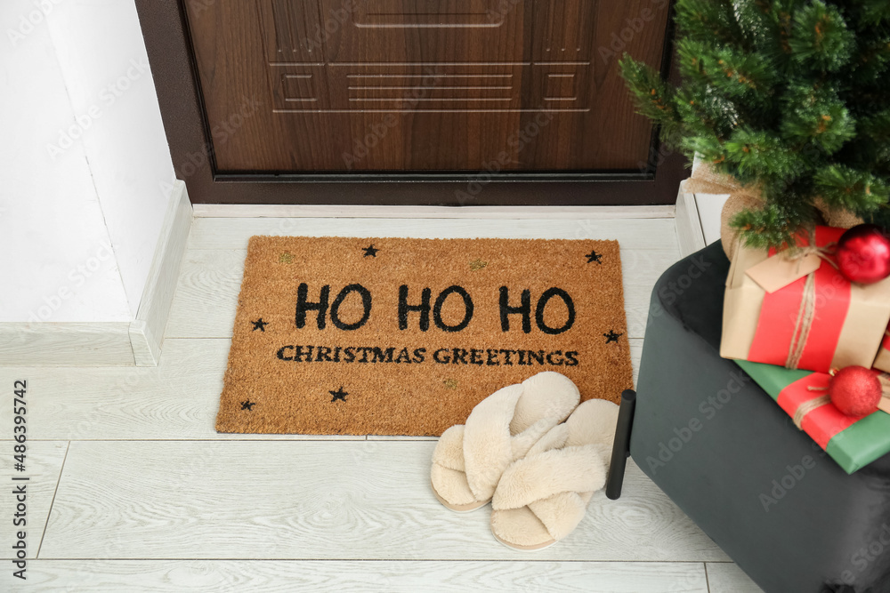 Mat with Christmas greeting and slippers near dark wooden door