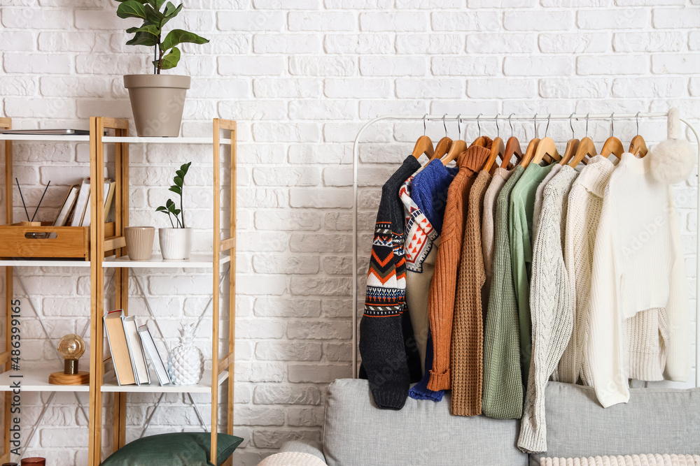 Shelf unit, sofa and rack with sweaters near white brick wall