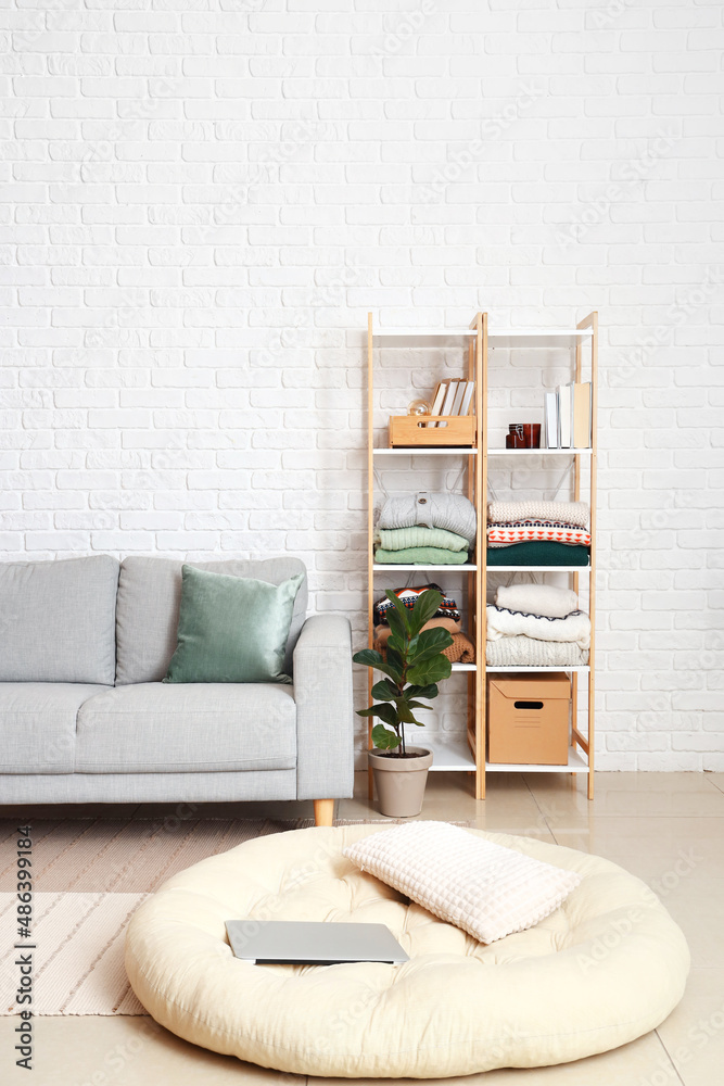 Comfortable sofa and shelf unit with folded sweaters near white brick wall