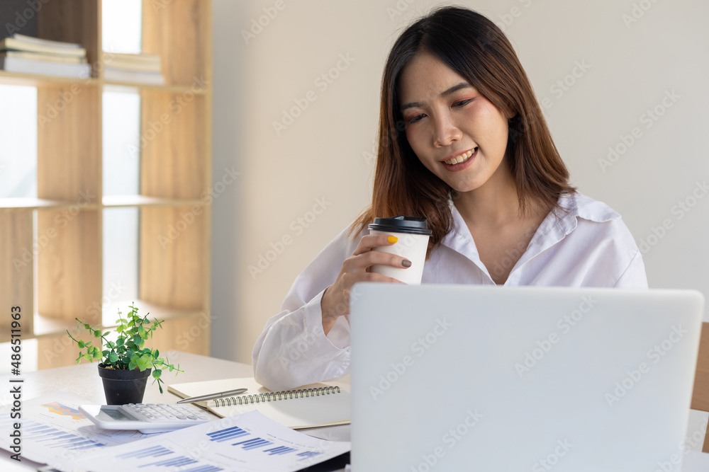 Asian woman in office casually drinking coffee working with laptop on her desk.