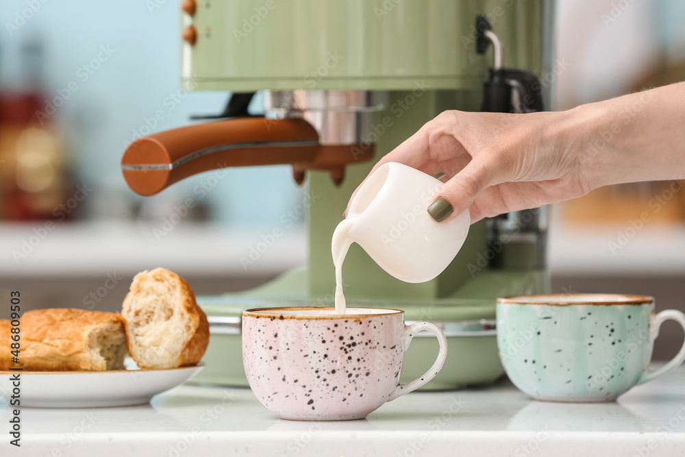 Woman pouring milk into cup of fresh coffee on table in kitchen