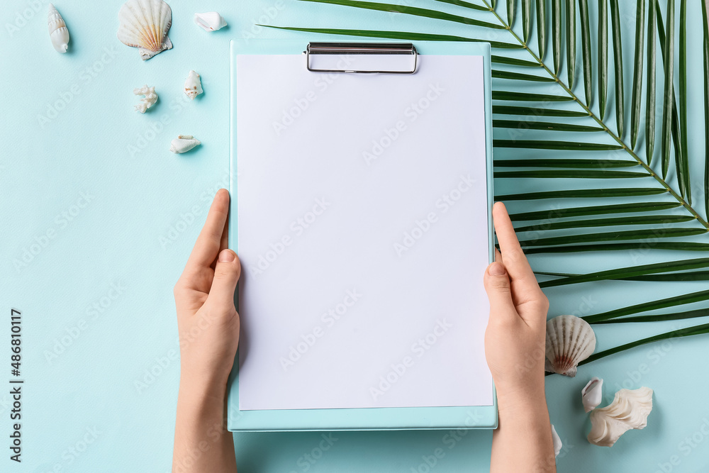 Female hands with clipboard, seashells and palm leaf on color background, closeup