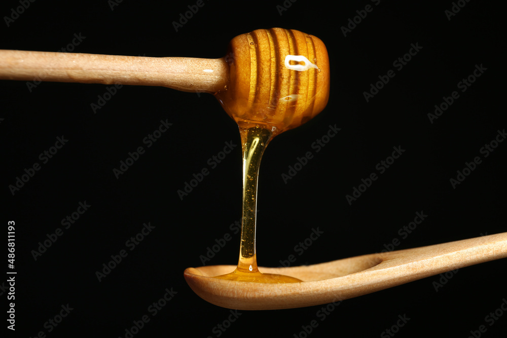 Honey dripping from dipper into wooden spoon on black background, closeup