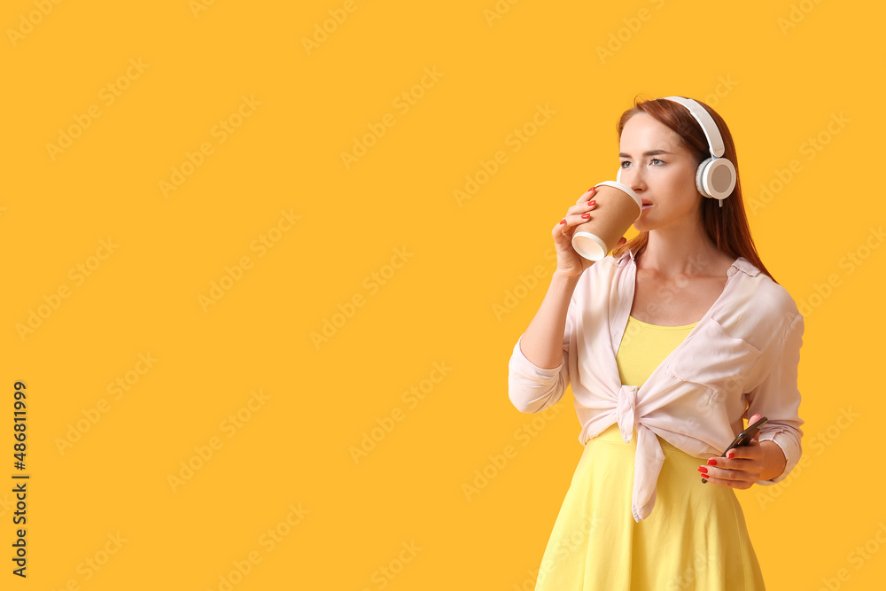 Young redhead woman in headphones with mobile phone drinking coffee on yellow background