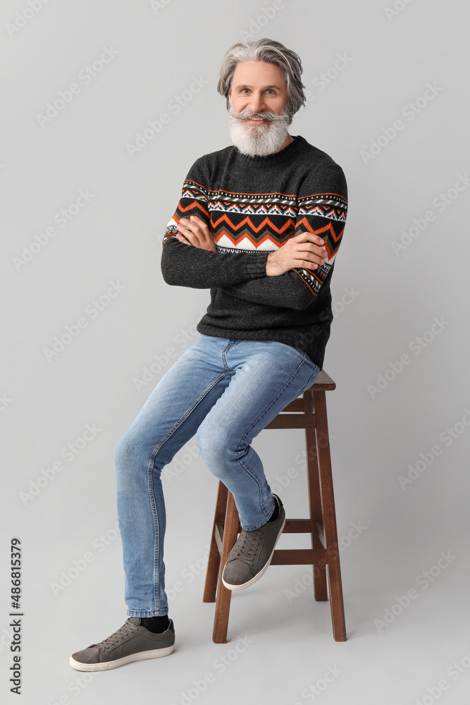 Handsome senior man in knitted sweater sitting on chair against grey background