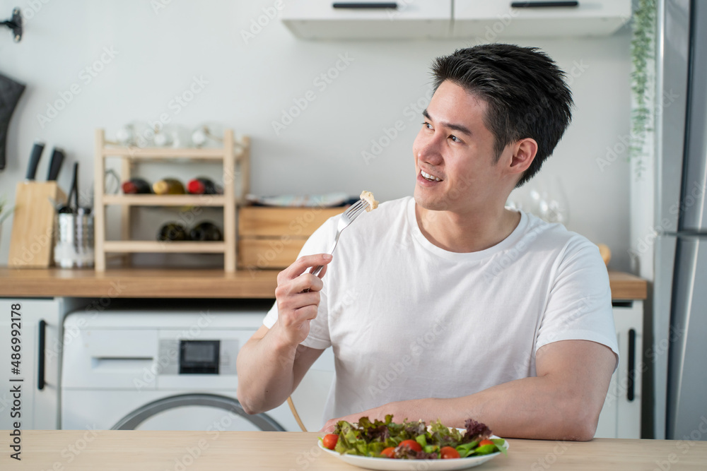 Asian attractive handsome male eating green salad in kitchen at home. 