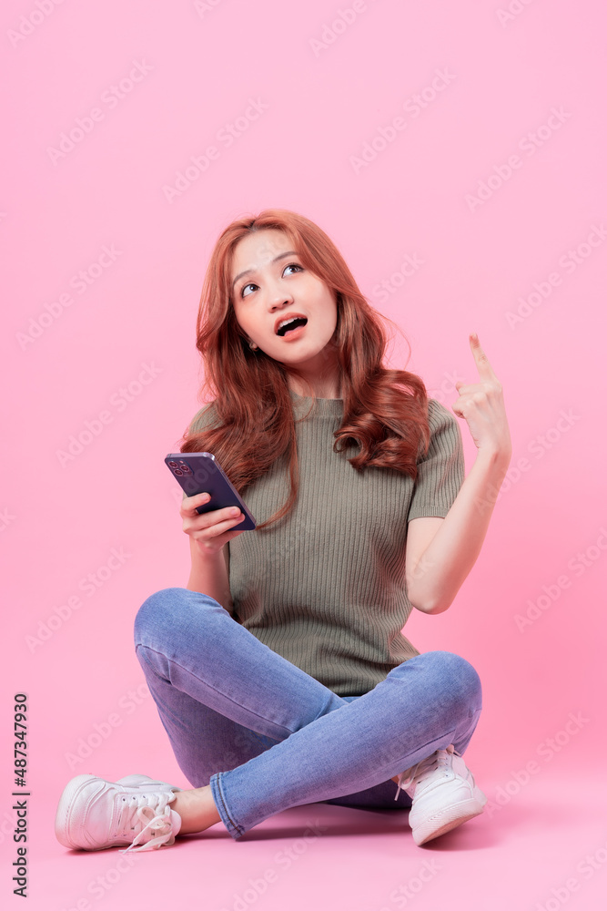 Young Asian woman sitting and using smartphone on pink background