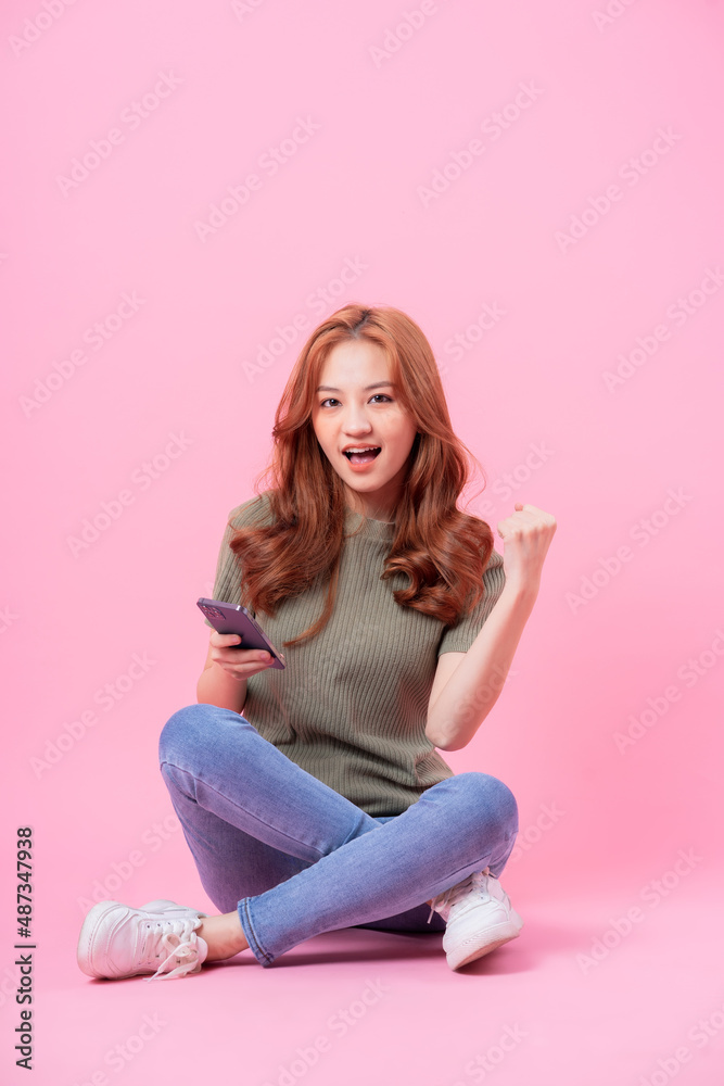 Young Asian woman sitting and using smartphone on pink background