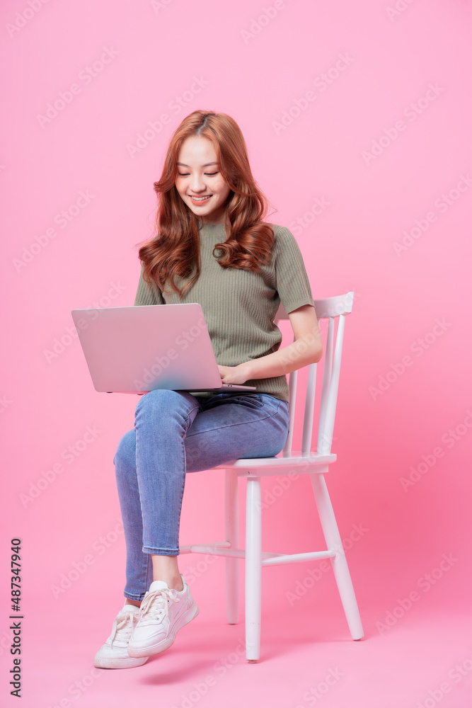 Young Asian woman sitting and using laptop on pink background