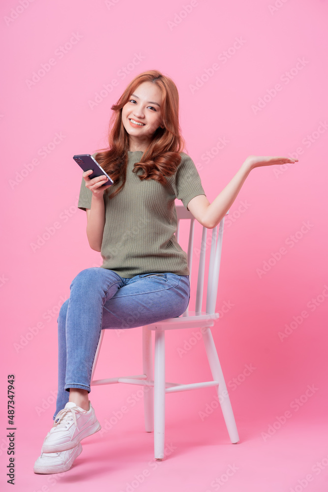 Young Asian woman sitting and using smartphone on pink background