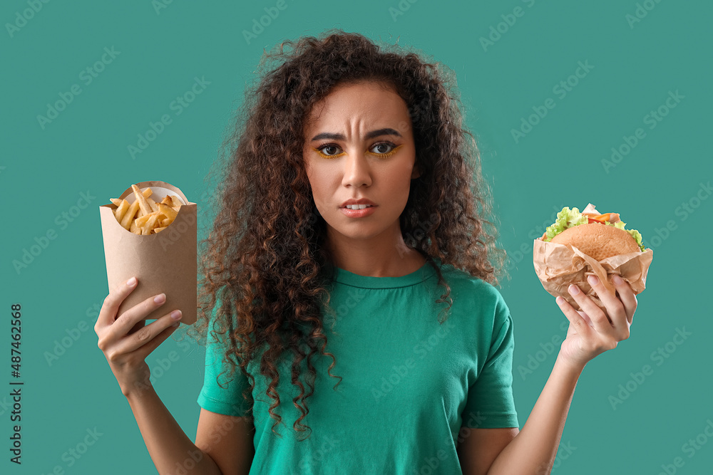 Sad young African-American woman with french fries and burger on color background