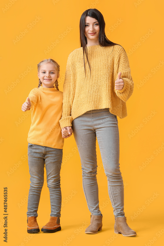 Little girl and her mother in warm sweaters showing thumb-up on yellow background