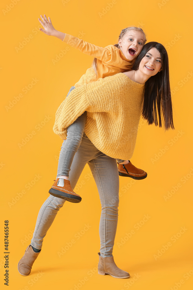 Young mother carrying her little daughter piggyback on yellow background