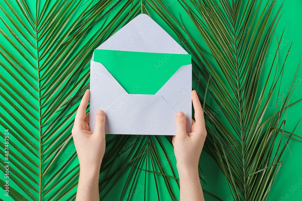 Female hands with card, envelope and palm leaves on green background