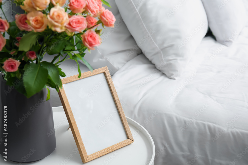 Vase with bouquet of beautiful fresh roses and blank frame on table in bedroom, closeup