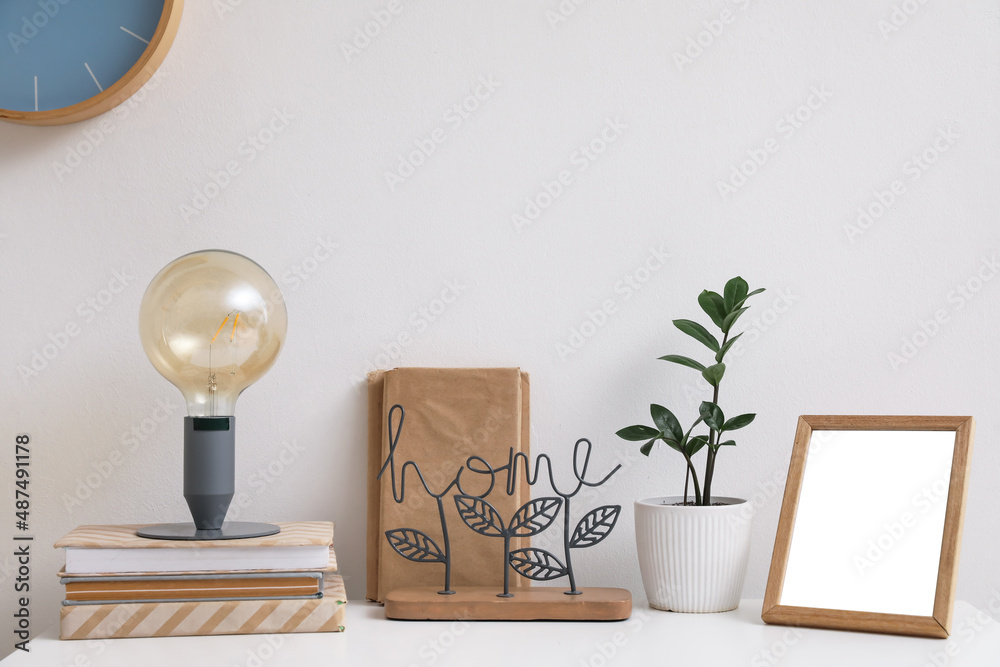 Stylish lamp, books, frame and flowerpot on table near light wall