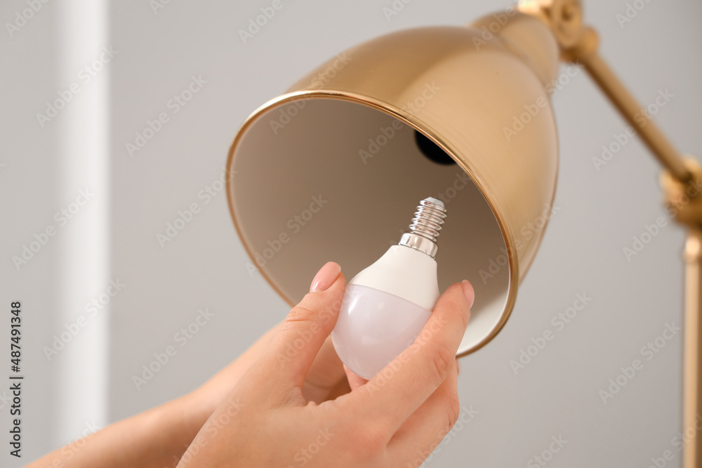 Woman changing light bulb in golden lamp near light wall, closeup