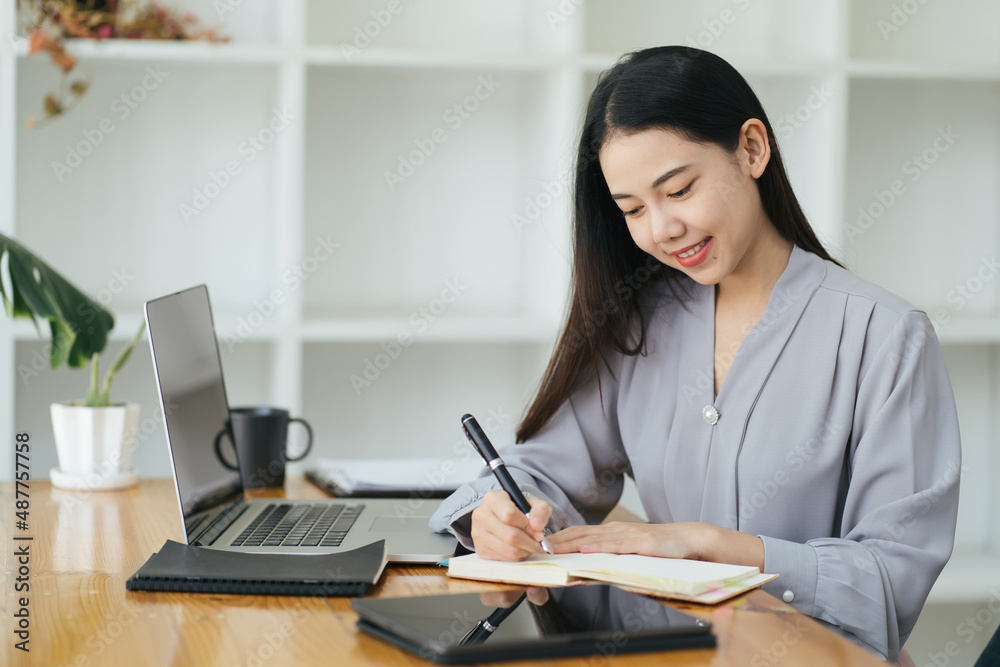 年轻女商人坐在办公室的电脑前，在笔记本上做笔记的肖像。