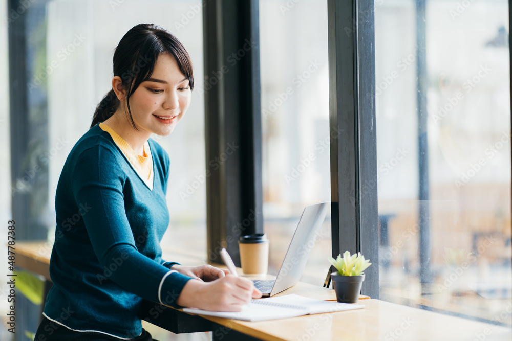 Young Asian woman browsing laptop while taking notes in planner sitting at table with laptop and cup