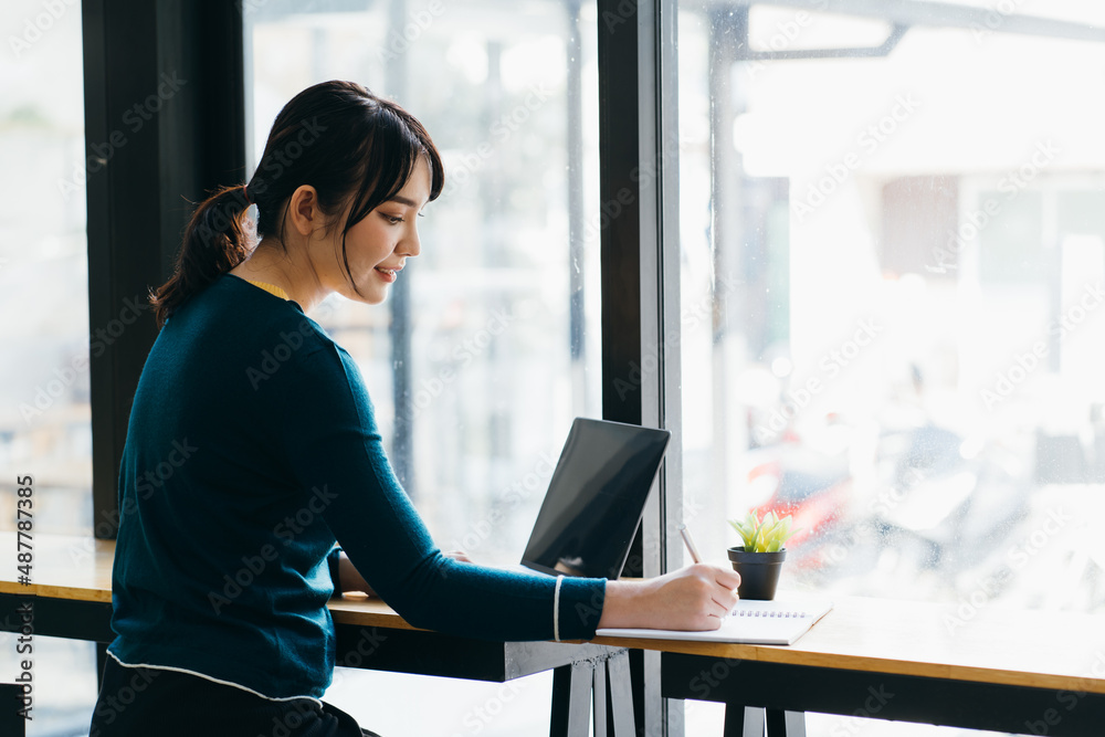 Young Asian woman browsing laptop while taking notes in planner sitting at table with laptop and cup