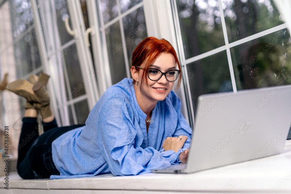 A woman is smiling and uses laptop while lying across the wooden platform. Working from home in quar