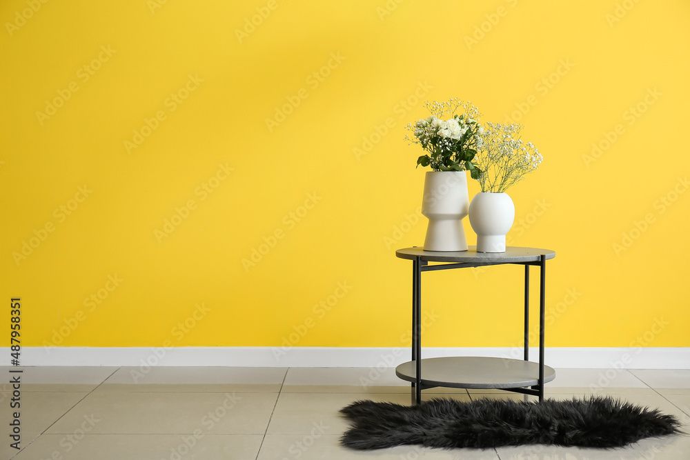 Vases with roses and gypsophila flowers on table near yellow wall