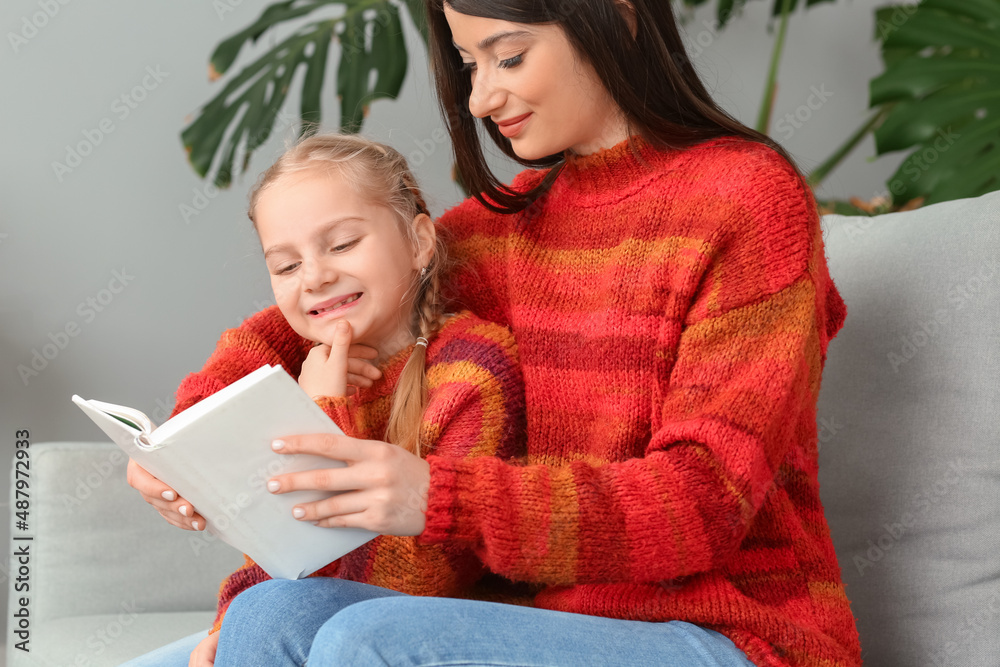 Little girl and her mother in warm sweaters reading book on sofa at home