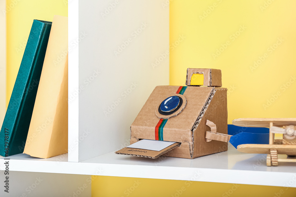 Photo camera made of cardboard on shelf unit near color wall